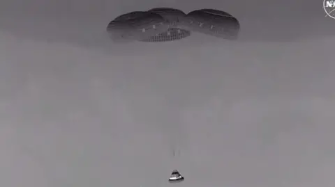 Black and white picture of capsule being hanging below three parachutes