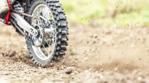 The back wheel of a scrambler motorcycle being driven across mud. 