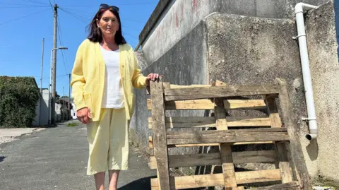 A woman is standing on a back street beside a wooden pallet that is leaning against a concrete wall. She is dressed in a yellow cardigan with a white top and yellow trousers. 