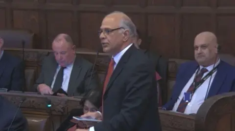 Coventry City Council Pervez Ahktar stands in the wooden panelled council chamber speaking into a microphone surrounded by colleagues seated on wooden benched. He wears a black suit jacket, white shirt and red tie.