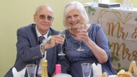A smartly dressed man and woman  smile and hold up champagne glasses in celebration