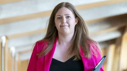 PA Media Gillian MacKay walking through a corridor in Holyrood - she has brunette hair past her shoulders, a pink jacket on and a folder or possibly a closed laptop in one hand 