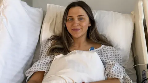 Ami Clarke Ami Clarke in a hospital bed wearing a gown. She has long dark hair and is smiling at the camera.