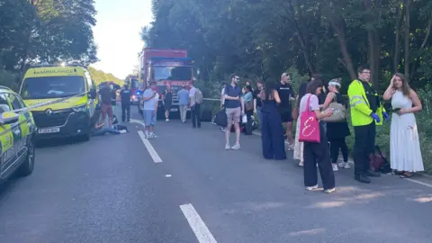Dr Peter Knapp A large group of people standing in a road, with emergency vehicles around them. Most look shocked and some have bandages and blood on them. 