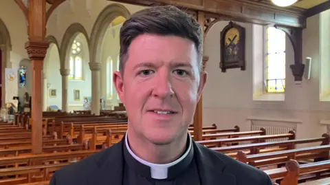 Fr Micheál McGavigan's mouth is partly open in a half-smile. His thick brown hair is lifted at the front and swept back to the right. He is wearing a clerical suit and collar. Behind him are rows of hazel pews, stained glass windows and iconography. 