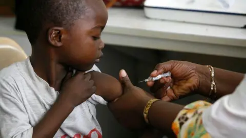 A young African boy is looking down at his arm as he is about to receive an  injection