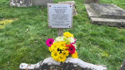 A close-up of a 17th-century grave with an inscription and a small bouquet of yellow and pink flowers on it.