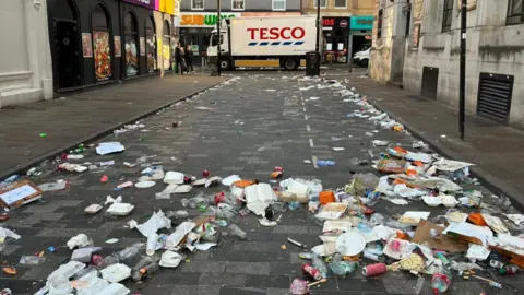 A white Tesco-branded lorry is the focal point of the photo, with a litter-covered road leading up to the lorry. Litter includes food boxes, plastic cups and bottles