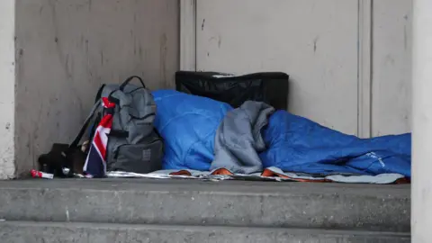 PA Media A rough sleeper in a blue sleeping bag on the ground with his grey back next to it 
