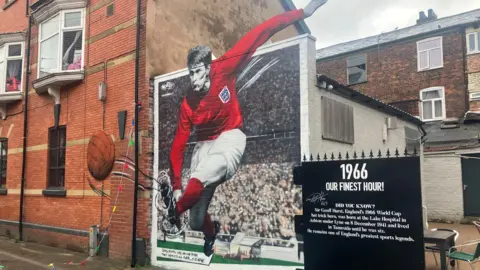 Photograph of the mural of Sir Geoff Hurst in Ashton-under-Lyne. The image shows the painting on the wall of a bar on Wood Street. Sir Geoff is wearing a red England shirt and kicking an old brown football, which appears to be coming out of the wall.