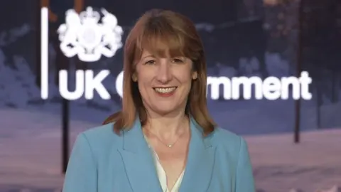 Rachel Reeves dans un costume bleu clair avec une chemise en soie blanche et un collier en or. Elle a des cheveux marron / cheveux bruns / rouges. Elle sourit devant un écran appelé le gouvernement britannique.