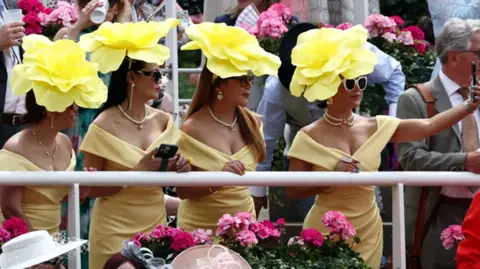 Reuters Four women, wearing yellow hats that look like flowers, lined up, with the woman on the right hand side taking a selfie of them on a mobile phone.