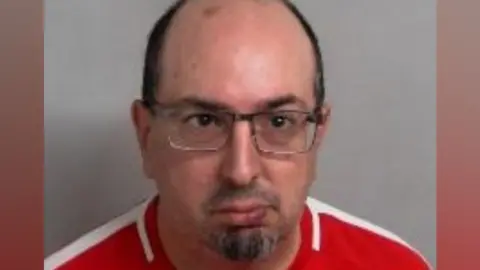 Essex Police Adam Evans pictured in police custody. He has a shaved head, a goatee and is wearing a red Nike top and glasses.