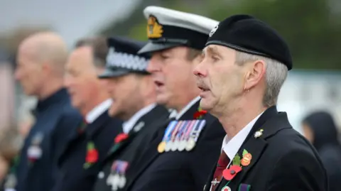 Penzance Council A line of men take part in the service. The four men are dressed in black uniforms. They are wearing poppies on their clothing as well as badges and medals. Their mouths are slightly open which suggests they are talking or singing. 