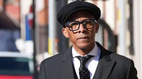 PA Media Image shows Jay Blades arriving at Worcester Crown Court wearing a black suit, white shirt, black tie with white spots and a tie pin and a red handkerchief tucked into his breast pocket. He is wearing a black peaked cap.