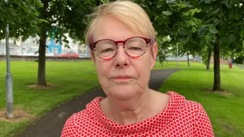 A woman with short blonde hair and purple highlights is wearing multicoloured framed glasses and is looking into the camera. She is wearing a red and white patterned top. She is standing on a path in a green area surrounded by trees.