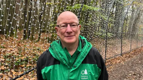 Jon Codd wearing a green Foresty England jacket, General Manager of Bedgebury England, stands in the Cathedral of Light installation with 39,000 fairylights 