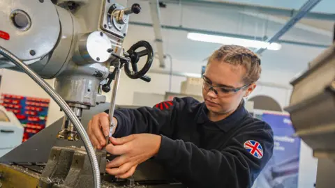 A woman wearing a jumper which has a union flag on the arm, is seen using a tool to drill downwards into some material. She is looking at her job.