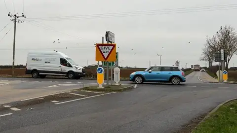 Looking across a four-way crossroads in a rural area as a white van and a blue car pass under a grey sky. A give-way sign and other signs stand in the centre of the picture. A lorry and a second car are in the distance. The land is flat with a brown field and bare trees, with birds sitting above on power lines.