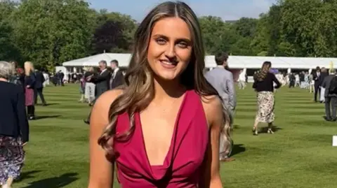 Zoe Walastyan standing in a garden at Buckingham Palace, with people and a marquee behind her. She has long fair hair and is wearing a red dress. 