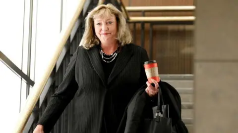 Marilyn Gladu walks down a staircase in a black suit with a silver necklace. She is holding a red takeout coffee cup in her hand as well as a black purse.
