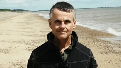 John Fairhall/BBC Dave Thurlow standing on a sandy beach strewn with pebbles at the sea edge on his right and behind him. He has short greying dark hair and is wearing a black jacket. 