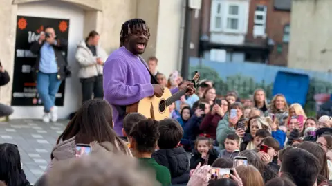 Myles Smith wearing a purple jumper and singing while playing guitar to a gathered crowd on a Luton street.