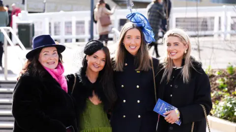 PA Media Four women, three of them young and one slightly older, smile as they stand arm in arm at the Cheltenham Festival. They are all wearing smart dark jackets and hats or fascinators