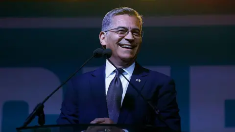  Xavier Becerra is seen on stage wearing a suit. He is smiling 