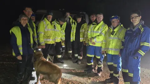 Swanage Coastguard A group of people from HM Coastguard stand around at night in a posed photograph. Uska, a golden-coloured Labrador, is standing in the centre, looking up at one of the men. 