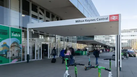 Milton Keynes station seen from the front entrance. It is a glass windowed building with a cover on pillars labelled Milton Keynes Central - with the National Rail logo. LOts of people are milling around on paving outside the station and taxis line up in the distance. 