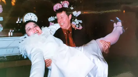 Handout A smiling woman in a white dress, white blazer and black shoes is being physically carried by a grinning man with dark brown hair.