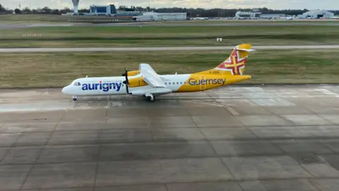 BBC An Aurigny aircraft on an airport runway. The plane is yellow and white and has two propellers.