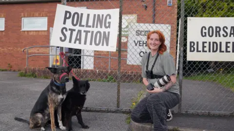 Photographer Ellen Cooper outside a polling station with two dogs sitting on the ground next to her. Both are tied to a chainlink fence with a red lead. Ellen has dark, animal print trousers on and a grey T-shirt. She is carrying a camera in her hands and has a tattooed left forearm and orange hair. A Polling Station sign hangs above the dogs and the building in the background is red brick 