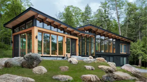 An exterior image of the Barlochan cottage. It is surrounded by greenery and rocks. It is a wooden structure that features the natural finish of Douglas fir and black paint on itse exterior. It is almost entirely exposed with floor-to-ceiling windows, and has two slanted roofs side-by-side. 