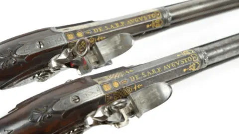 Two decorative 18th Century pistols against a white background with the words "DE. S.A.R.P. AUGUSTO" written on them in golden gilt.
