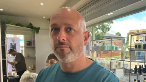 Adele Forrest/BBC A man with a short white beard and bald head in a blue T shirt stands in a hair salon