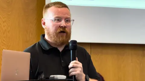 Applause Business Consultancy Rob Smith holds a microphone and speaks at the event. He has red hair and a beard, wears glasses and a black polo shirt. 