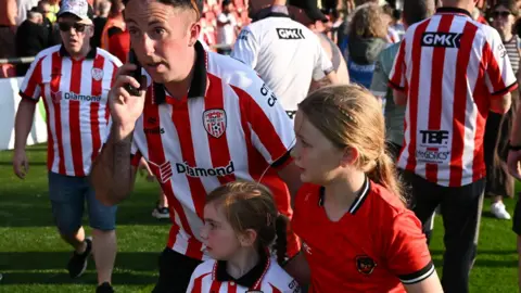 Getty Images Two girls in Derry City tops look fearfully out of frame as a young man with them talks on the phone. There are crows of people around them, standing on a football pitch
