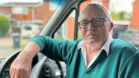Head and shoulders image of Dave Shepherd in the bus driver seat. He has short white hair, dark rimmed glasses and is wearing a green jumper with a white shirt. He has his right arm on the steering wheel.