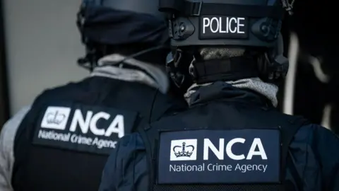 Two National Crime Agency (NCA) officers pictured from behind. They are wearing black jackets and NCA bulletproof vests, as well as black helmets.