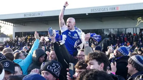 Macclesfield's Josh Kay raises his arm in the air celebrating while surrounded by fans on the pitch. His mouth is open and he looks to be cheering. He appears to be on the shoulders of a team-mate and raised above the surrounding fans.