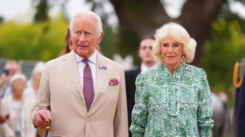 King Charles III in a tan suit and purple tie. Queen Camilla is standing next to him in a green dress.