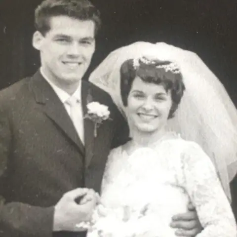 Supplied A wedding picture of Frank wearing a dark suit shirt, tie and flower pinned to his jacket. He has his arm around his bride, Cath who is wearing a white wedding dress with a veil and holding a bouquet of flowers. 