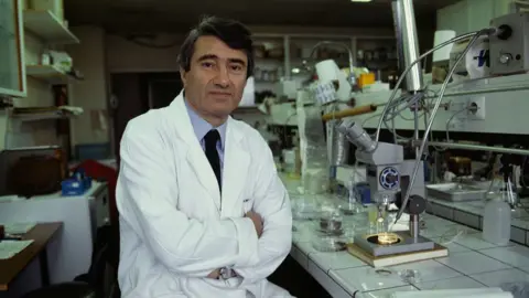 Dr Etienne-Emile Baulieu sits in a white lab coat with his arms crossed over his waist inside a lab, with various equipment around him and in the background