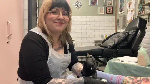 Kim Thomas in her studio smiling at the camera, holding a needle and wearing black gloves and a plastic apron with a customer's tattooed legs visible in front of her 