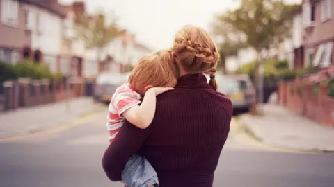 Getty Images Rear view of a woman carrying a child in a street. Their faces are hidden.