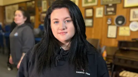 Emma Lewis, black long hair wearing a black hoodie with a Counter Force logo.