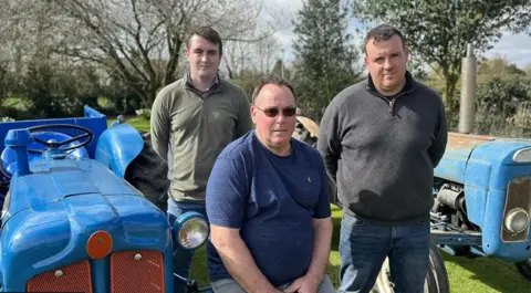 The father sits in the foreground and the sons stand behind him. Two blue tractors are parked either side of them. One has a rusted bonnet.