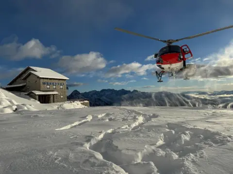 Soccorso Alpino e Speleologico Trentino Red helicopter with white underbelly prepares to land on snow close to a lodge on a sunny day with blue sky and clouds